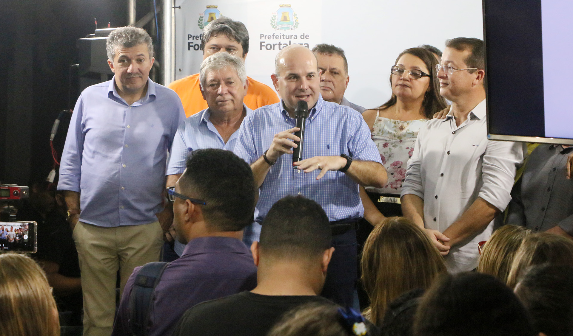 prefeito falando ao microfone em cima de palco com pessoas ao lado e outras assistindo à frente